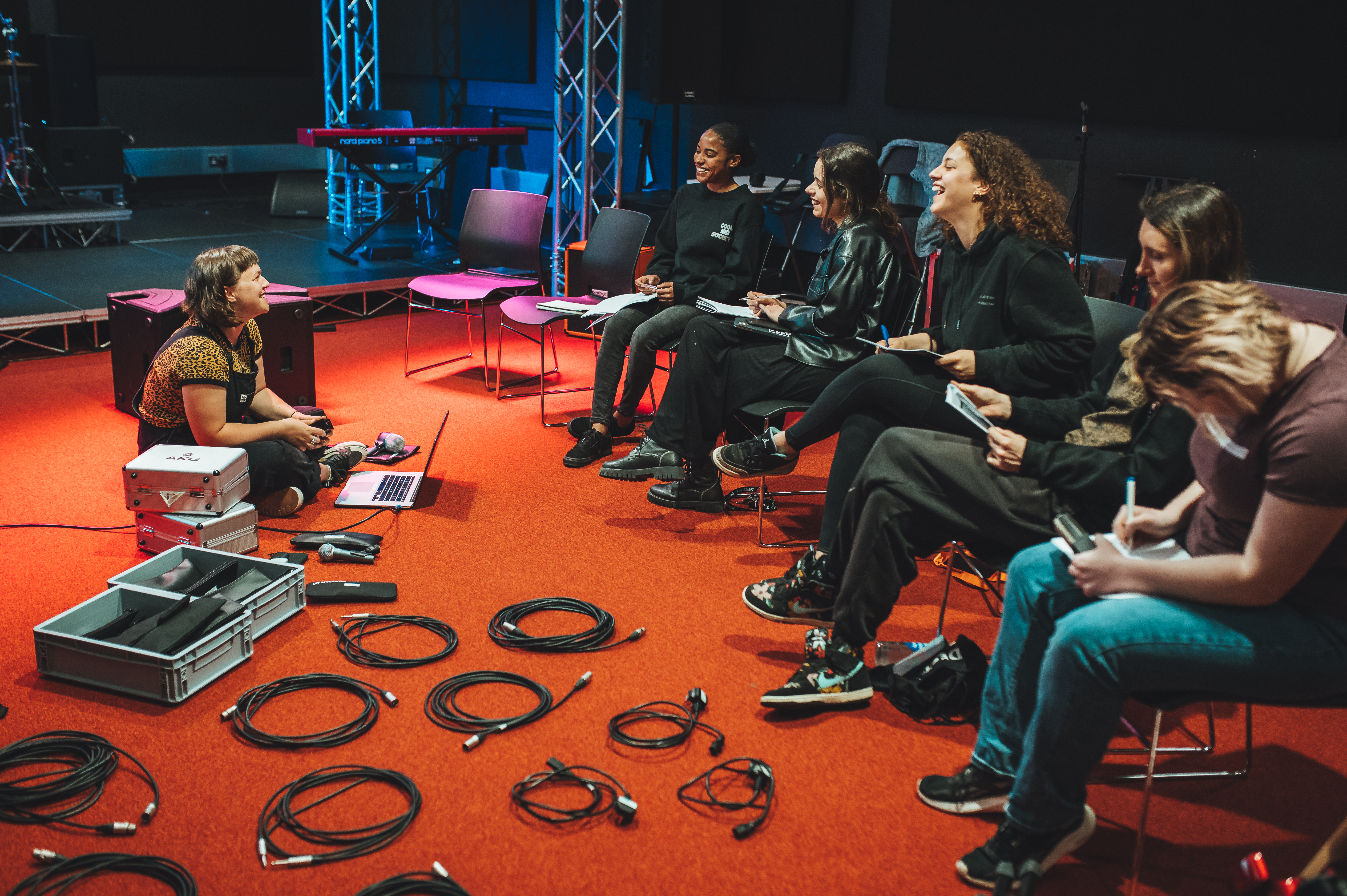 a group of women sit in a semi-circle on chairs, laughing. there are electric cables on the floor as one woman sitting on the floor in front leads the session 