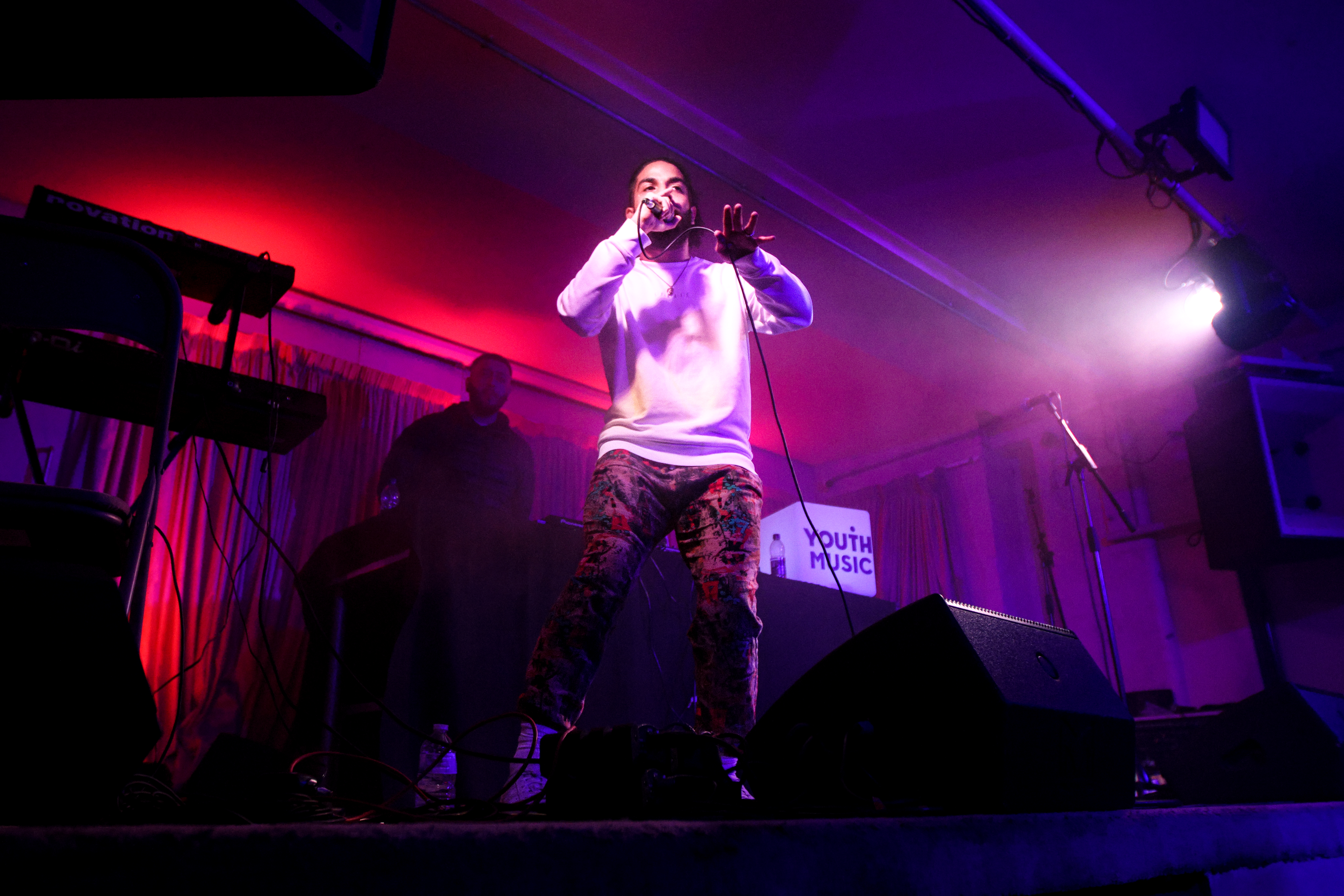 a man in a white jumper and patterned trousers stands on stage and sings into a microphone