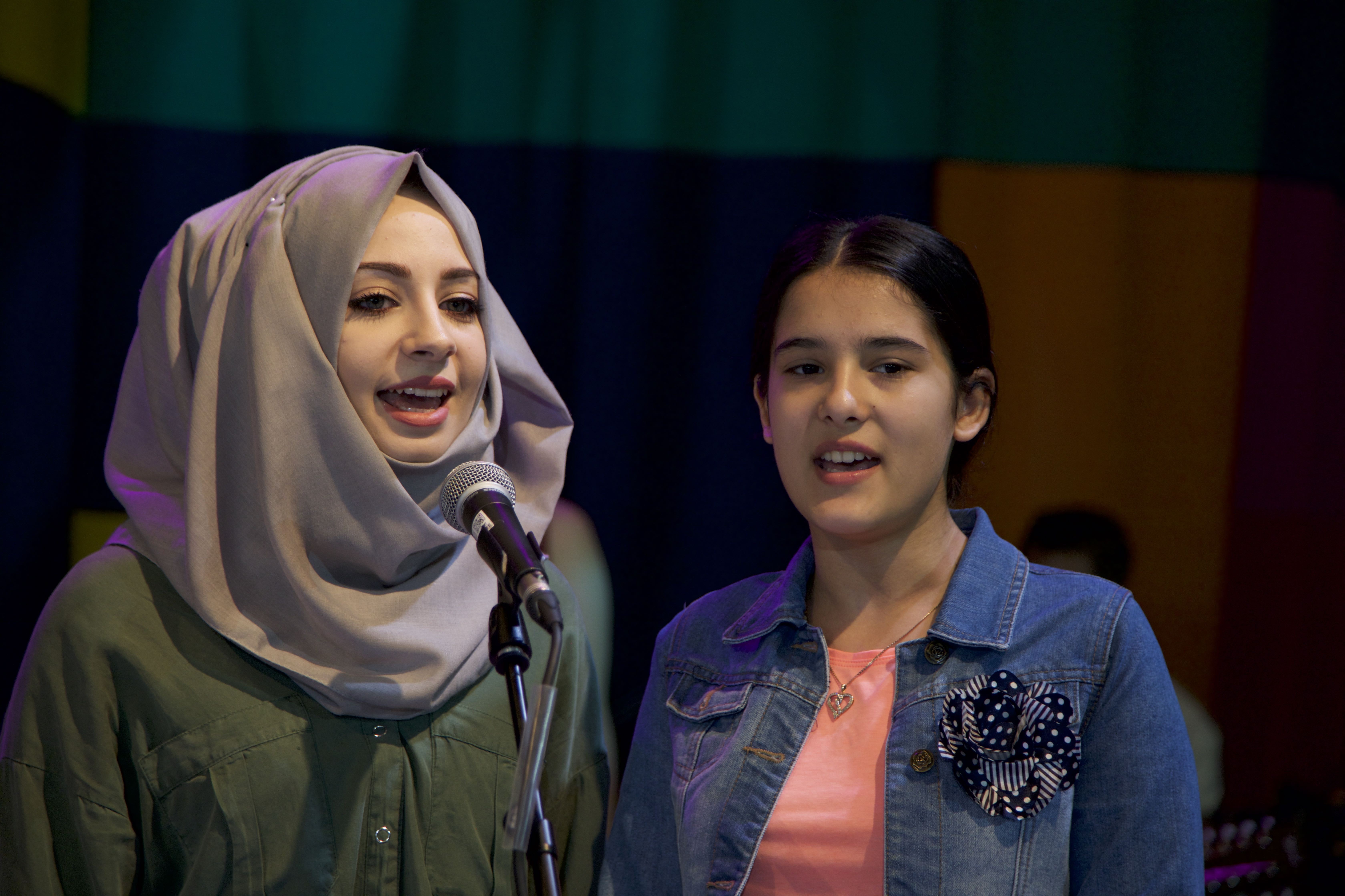 two girls sing; one in front of a microphone 