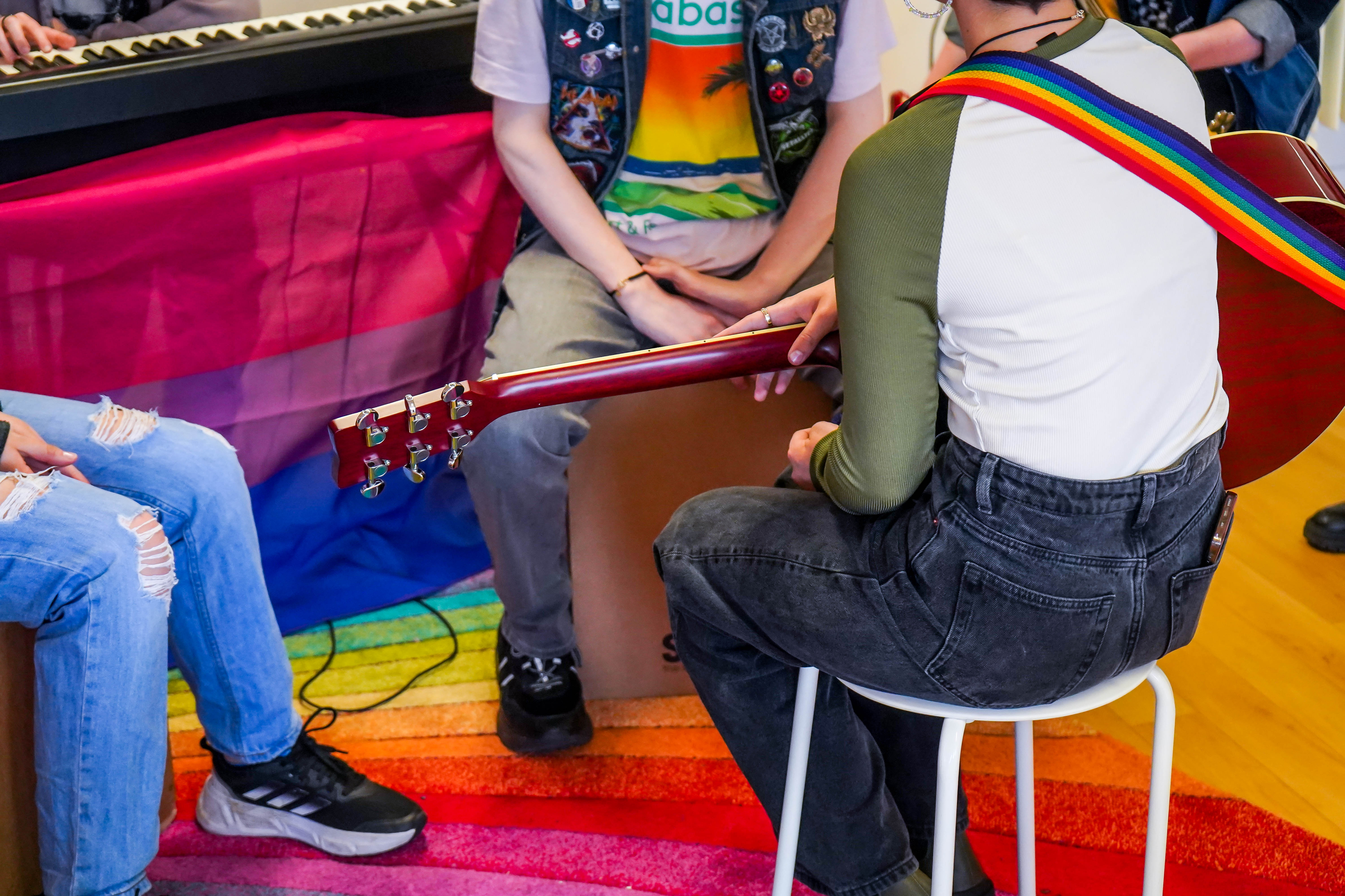 4 young people sat around a guitar with a rainbow guitar strap and rainbow carpet