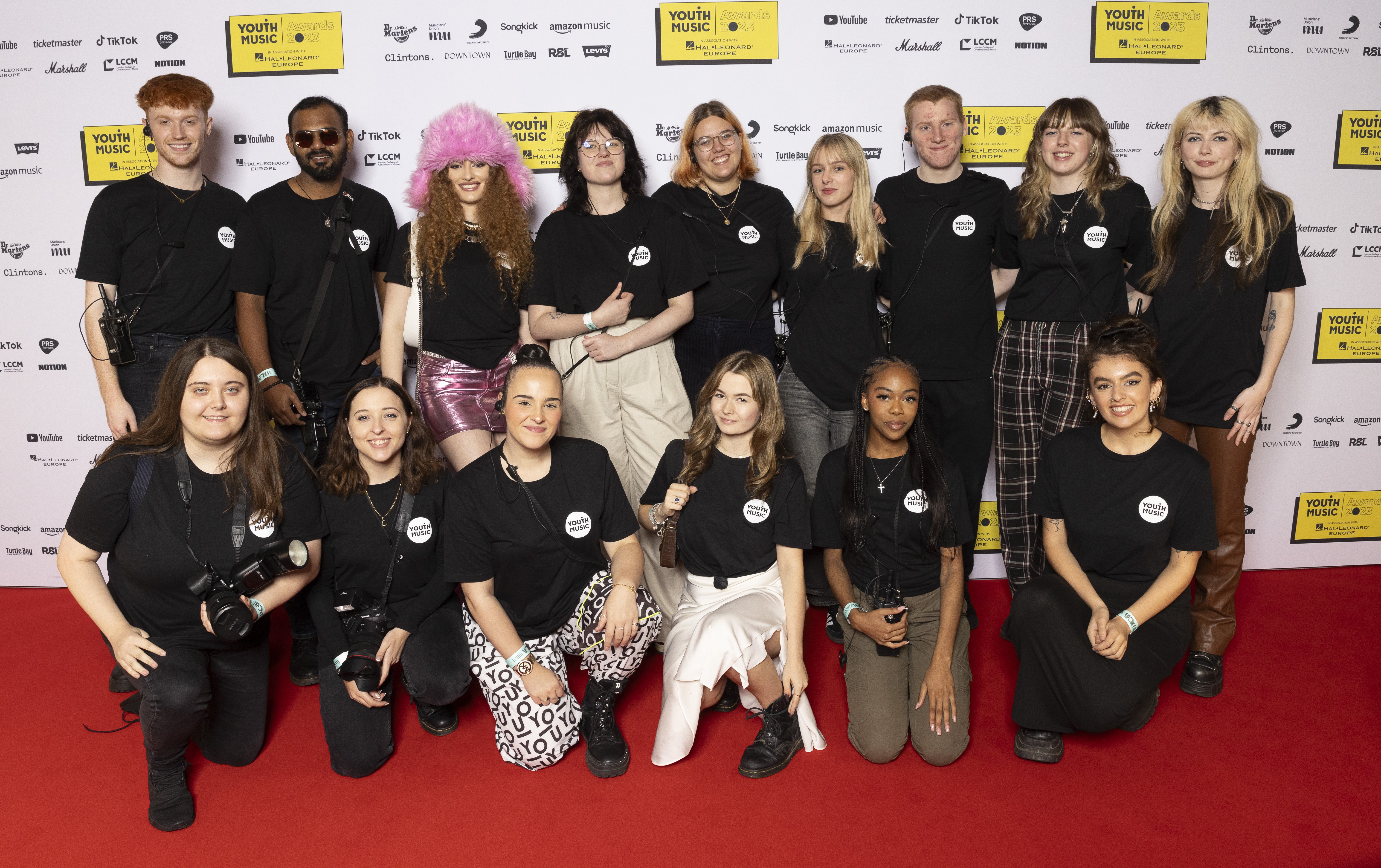 a group of young people wearing black youth music logo t-shirts pose together on a red carpet