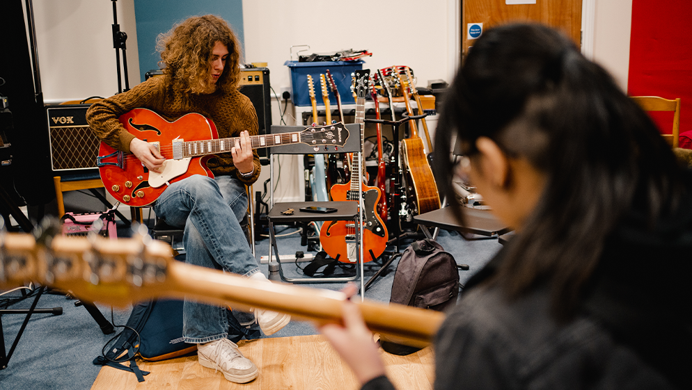 two people play guitars in a music room surrounded by more guitars