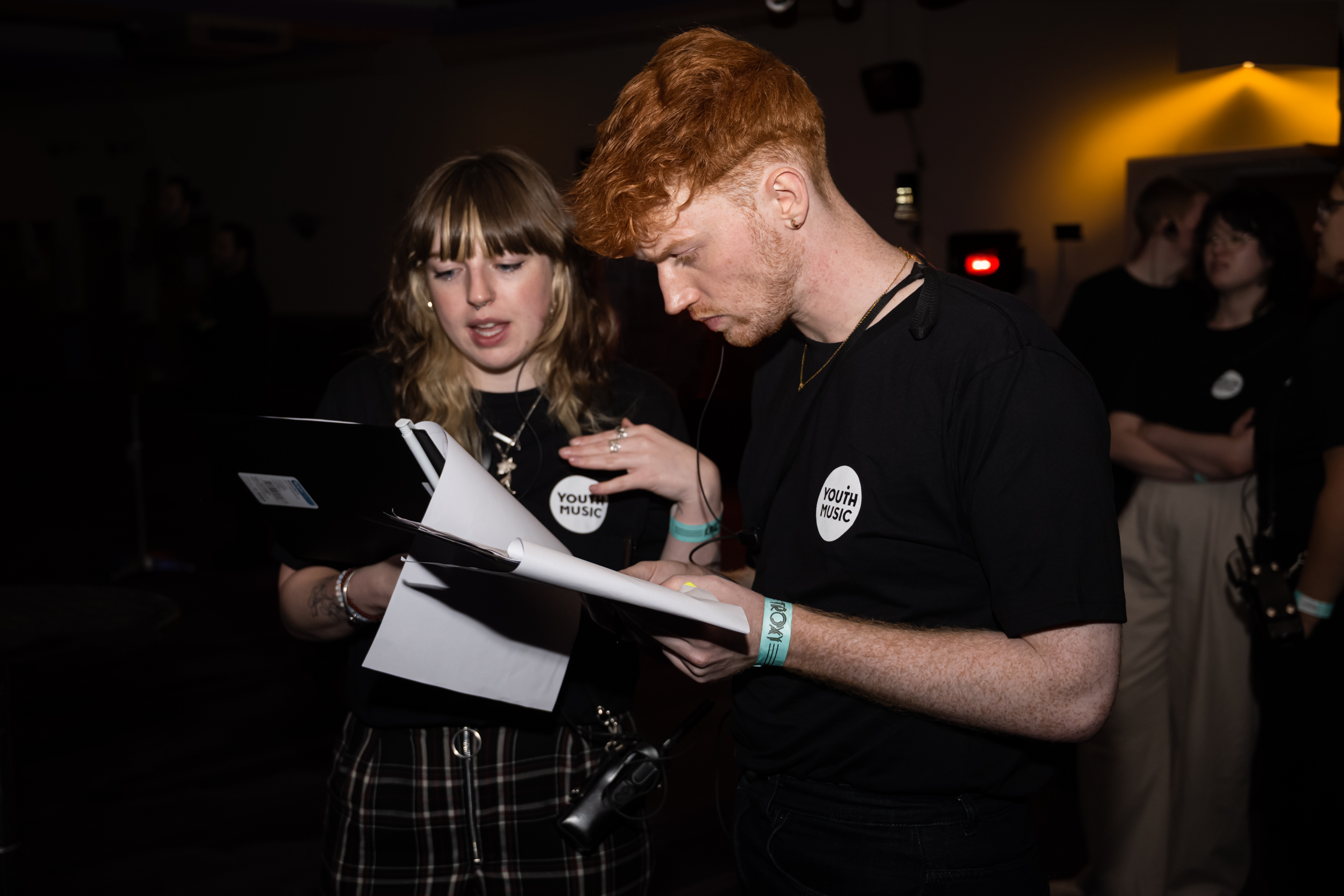 two young people look at clipboards in a dark room. they are both wearing black youth music awards t-shirts
