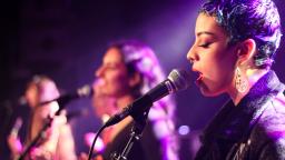 Live performance with three singers. Singer closest to the camera is in focus, and they have short purple hair, and wearing a black jacket and silver earrings