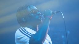 Young person wearing sunglasses and a white t-shirt with three black stripes, rapping on stage