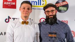 Two men in front of logo board, one with beard and glasses holding award trophy