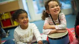 two children sitting on the floor, one with their arms on top of a drum, smiling at someone off camera