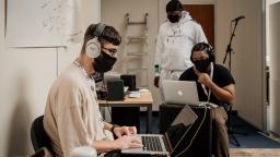 three people in a room working on laptops, all of them wearing black facemasks