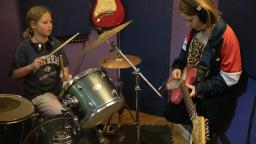 two young girls play instruments, one plays the drums and one plays the electric guitar
