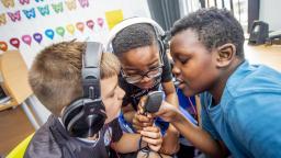 a group of children make music together. Two wear headphones and they all hold a microphone