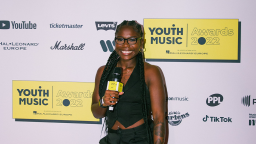jade barnett wears a black waistcoast and black trousers. her hair is in braids, she wears glasses and is smiling, holding a microphone