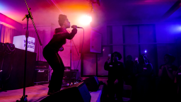 a woman sings on stage with ambient magenta lighting behind her. audience members hold up their phones to record her