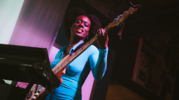 a Black woman wearing a blue long sleeve dress and blue eyeshadow plays a bass