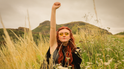 a woman with red hair stands in a field with long grass and raises her arm in the air, making a fist