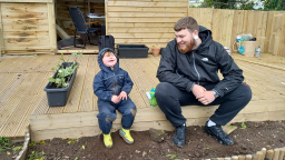 a dad with brown hair and a beard, and a toddler in a blue onesie sit on a wooden patio and laugh together