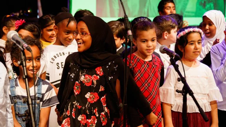 A group of children are singing and smiling on stage in front of three microphones. 