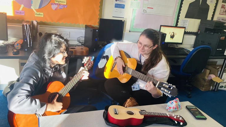 Two people sitting down playing acoustic guitars in a classroom.