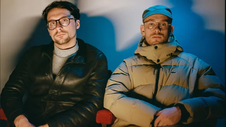 Harvey Gunn and Frankie Stew headshot, sat on chairs next to each other wearing coats