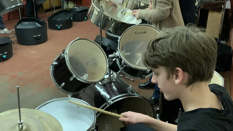 Young people in a music room. One is playing a drum kit and two others are playing steel drums.