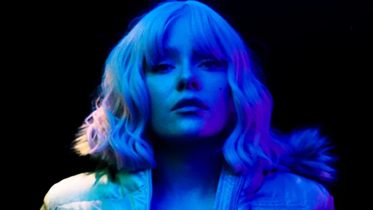 L C Y has a blonde wavy bob and is photographed under blue lighting