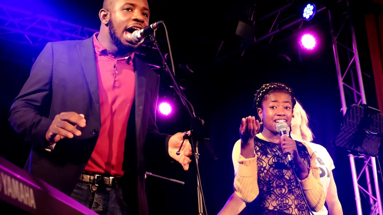 Two people on a stage singing into microphones, with red stage lighting behind them.