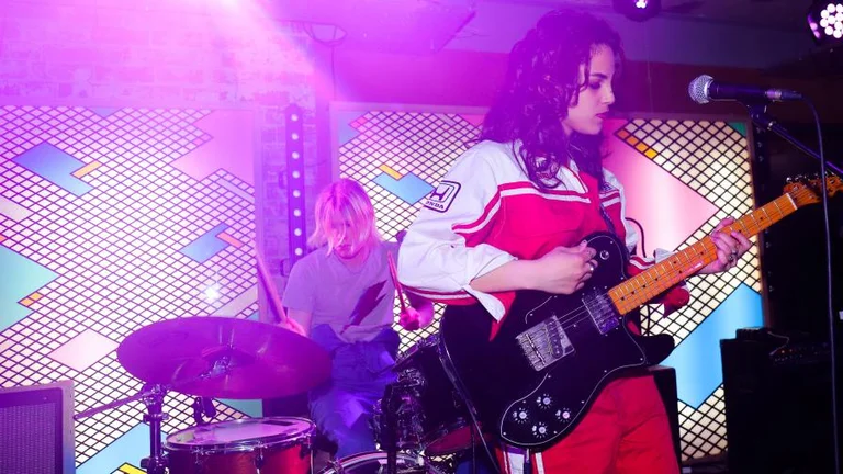 Young people performing on stage, one playing an black electric guitar, the other person people the drums