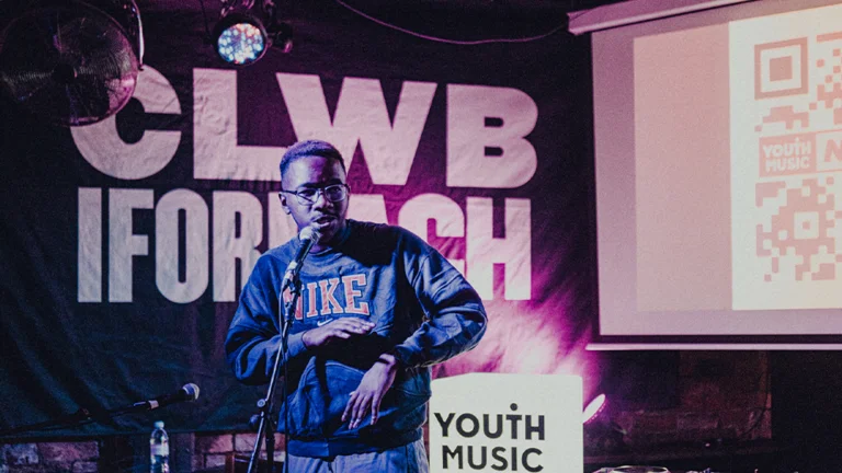 A performer on stage at a music venue in Wales. They are speaking into a microphone and there is a Youth Music logo behind them.