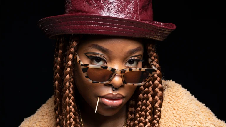 Meduulla, a woman with light braided hair, wearing a red leather hat, tortoiseshell glasses and a toothpick in her mouth.