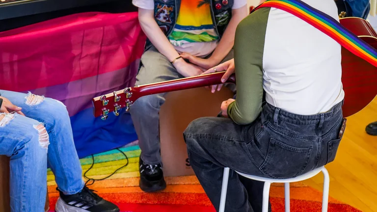 Young people with backs to the camera, rainbow carpet and rainbow guitar strap on an acoustic guitar