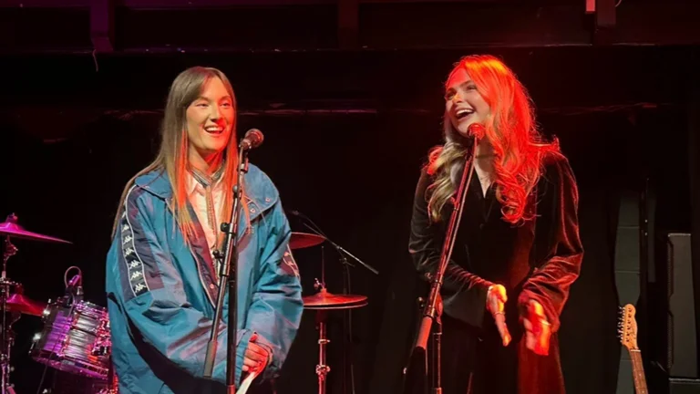 maria and sam stand on stage under red lighting, in front of microphones. they are laughing.