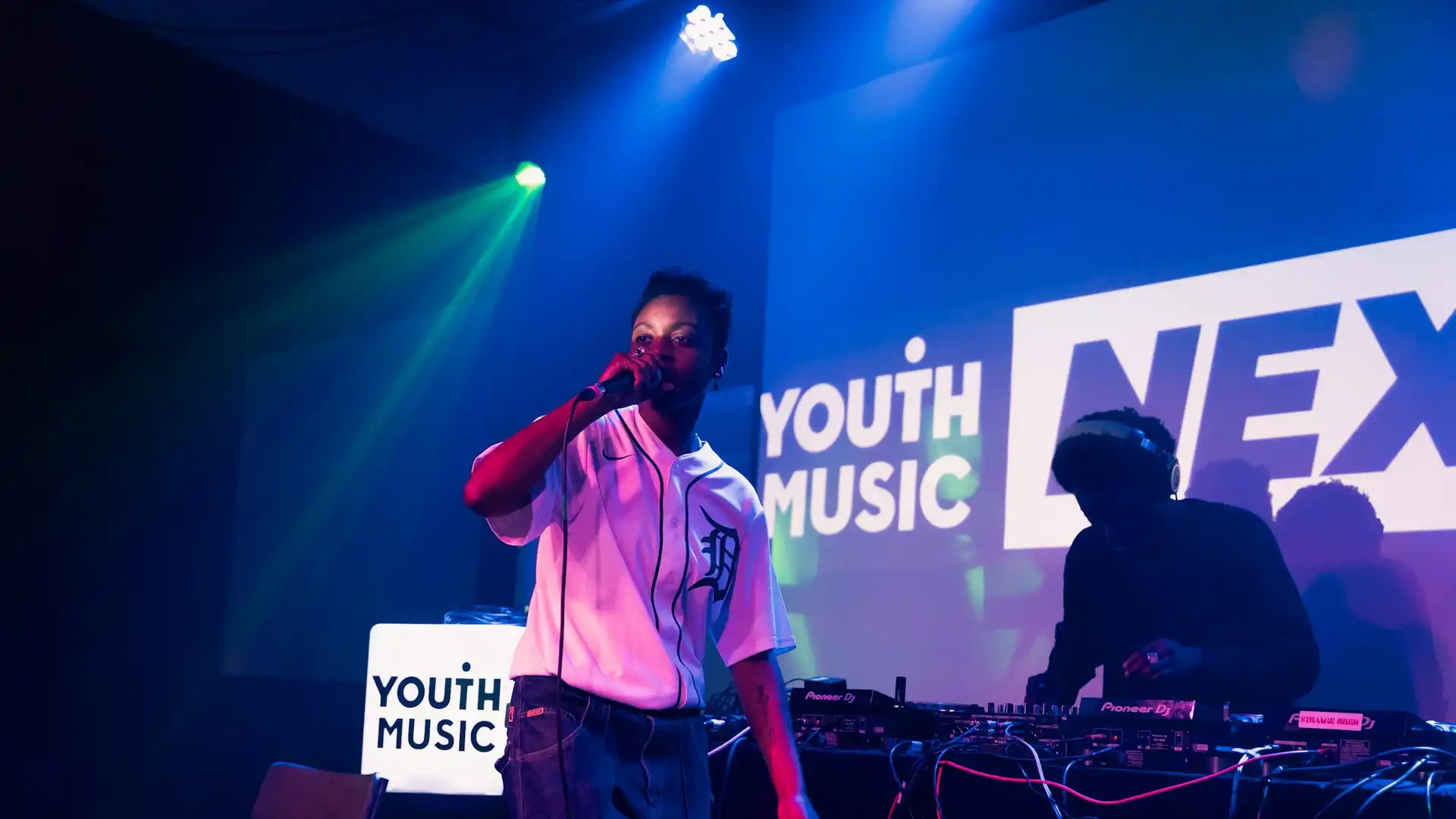 Black NextGen artist with a mic and DJ behind. The Youth Music logo is projected in the background. 