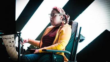 Person sitting in wheelchair wearing a yellow top. A black and white striped wall is behind them