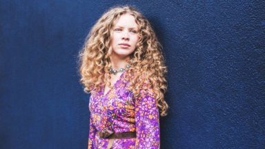 Image of Becca James. She has long curly hair, is wearing a purple, orange and pink floral dress, and is standing against a dark blue wall.