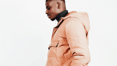 Image of Dami Sule. He is standing to the side, and wearing a beige hooded jacket over a black polo neck jumper.