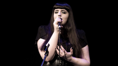 Image of IRIDIS performing. She has dark hair with a fringe, and is wearing all black clothing.