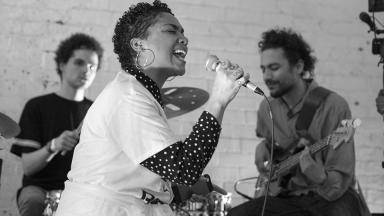 a woman sings into a microphone. two musicians are in the background. the photo is in black and white