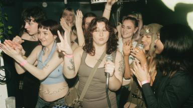 a group of young women cheer and clap in a room. one woman in the centre is holding a microphone