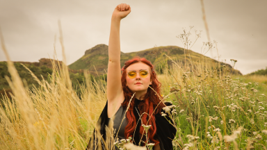 a woman with red hair stands in a field with long grass and raises her arm in the air, making a fist