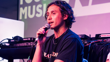 a man with brown curly hair speaks into a microphone. a dj set is behind him.