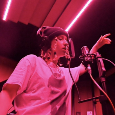 Young person with braids, wearing a black beanie and white top, singing in a studio.