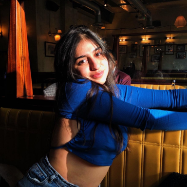 Photo of Prachee Mashru sitting in a booth with her arms stretched out, wearing a blue long sleeve crop top and jeans.