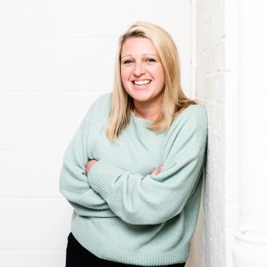 Woman with blonde hair wears green jumper and poses with folded arms