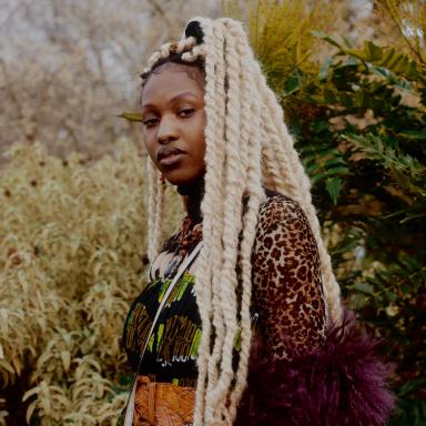 muva of Earth wearing multi-patterned outfit, stood outside in front of foliage