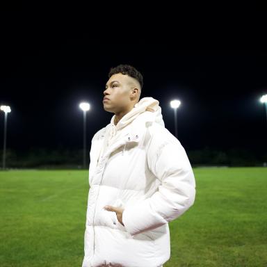 Keanan wearing a white coat, standing on a floodlit sports field