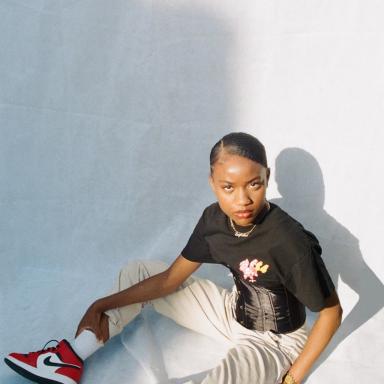 Rasida sitting down in front of white background, wearing black top, white jeans and red trainers
