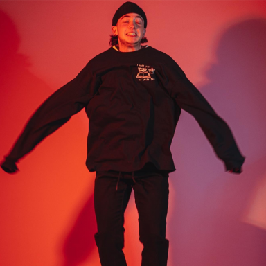 A press shot of Kai jumping in the air, with a atmospheric red background. He is wearing all black including a sweatshirt, jeans, boots and a beanie. 