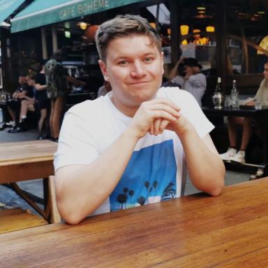 Iain sits at wooden bench smiling at camera wearing white t-shirt