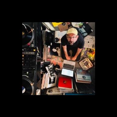 Chris sits on floor surrounded by vinyl records. He is wearing a black t-shirt, black rimmed glasses and a yellow baseball cap