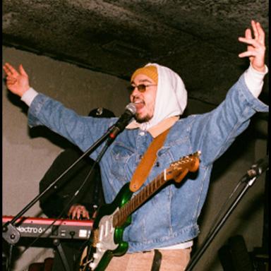 kalla wearing sunglasses, a denim jacket and white hoodie with the hood up. He is performing at a gig and wears an electric guitar around his neck