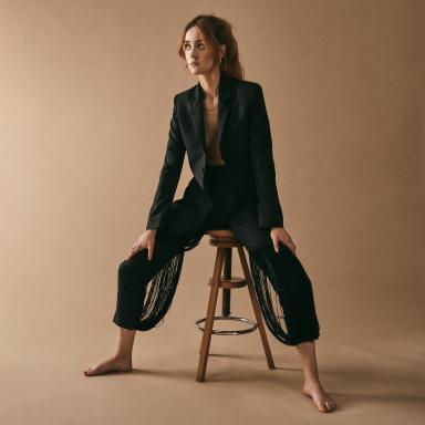 lees wears a black blazer and trousers as sits barefoot on a stool against a beige backdrop.